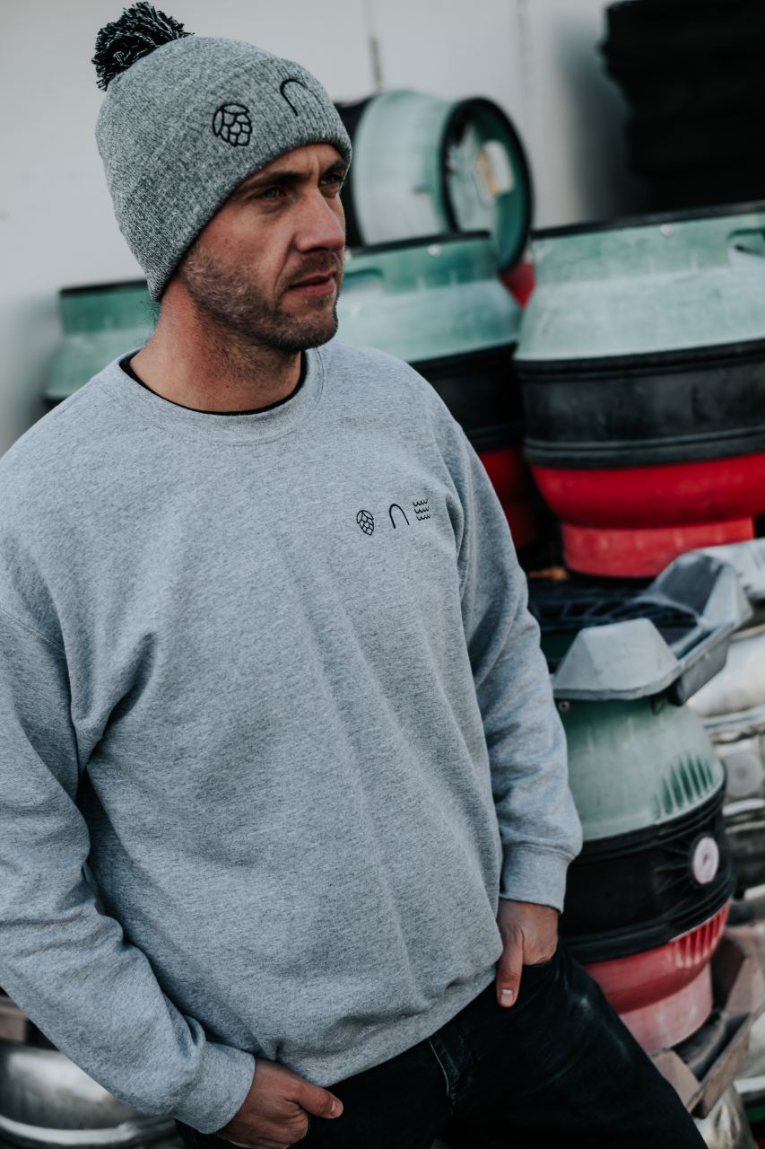 Man wearing a gray beanie and sweatshirt with a eightarch brewery logo, standing in front of beer kegs.